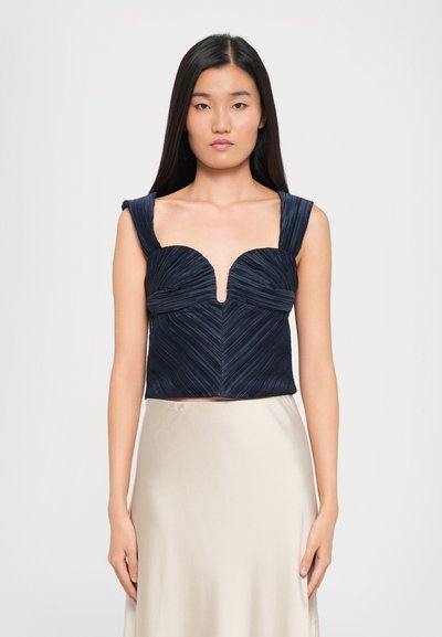 Top court bleu marine avec texture plissée, présentant un décolleté en V profond et des bretelles larges. Porté avec une jupe satin beige.