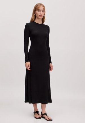 Zwarte jurk met lange mouwen van jersey stof, aansluitend aan de bovenkant, vloeiend tot midi-lengte. Draagt met zwarte platte sandalen, minimalistisch ontwerp, zonder patronen.