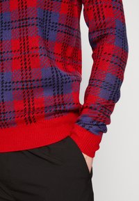 Rot und lila gestrickter Pullover mit klassischem Karomuster, gerippten Bündchen und Saum sowie einer weichen Textur. Kombiniert mit schwarzen Hosen.