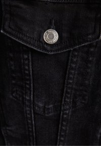 Black denim fabric features a metal snap button with engraved text. The pocket design includes stitched detailing and a smooth texture.
