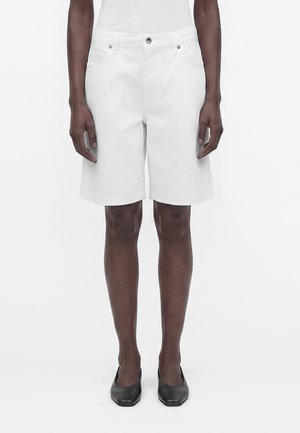 Person iført hvide knælange shorts, enkel hvid top og sorte slip-on sko, stående med armene afslappet langs siden.
