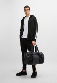 Felpa nera con strisce bianche, pantaloni neri attillati e sneakers nere, che tiene una borsa da viaggio nera con il logo "BOSS".