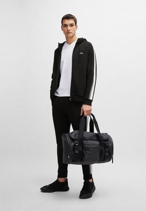 Haut à capuche noir avec des rayures blanches, pantalon noir ajusté et baskets noires, tenant un sac de sport noir avec le logo "BOSS".