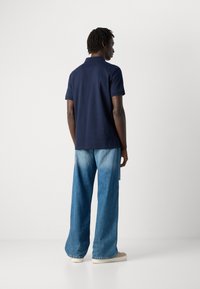 Férfi áll egy fehér fal előtt, sötétkék pólót, világoskék széles szárú farmernadrágot és bézs sportcipőt visel.