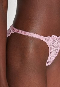 Krajkový tanga v jemně růžové barvě se květinovými vzory, vlnitými okraji a tenkým elastickým pasem. Hladká textura s jemnými detaily.
