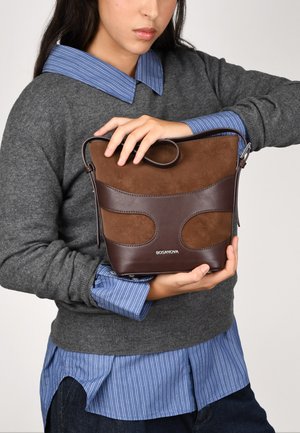 Bolso de mano de cuero marrón con detalles de ante, parte superior redondeada, correa y logo. Modelo sosteniendo el bolso mientras lleva un suéter gris y una camiseta a rayas.