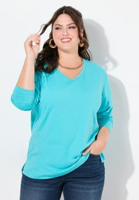 Tealfarbendes Langarmshirt mit V-Ausschnitt aus weichem Stoff, mit lockerem Schnitt und seitlichen Schlitzen, kombiniert mit dunkelblauen Jeans.
