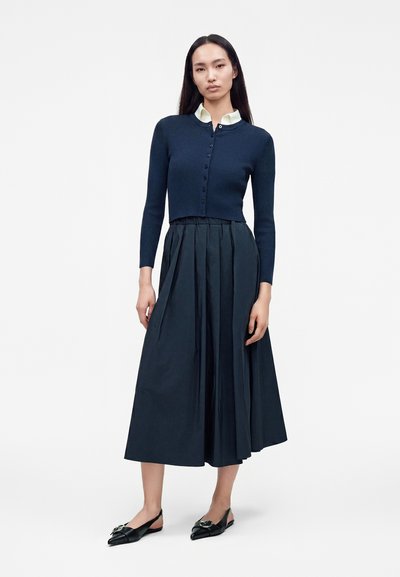 Femme portant un cardigan court bleu marine boutonné par-dessus une chemise blanche à col, avec une jupe midi plissée bleu marine et des ballerines noires pointues.