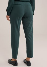 Zelené nohavice na mieru vyrobené z hladkej látky, s úzkym strihom, dvoma zadnými vreckami a rovnou nohavicou s manžetou.