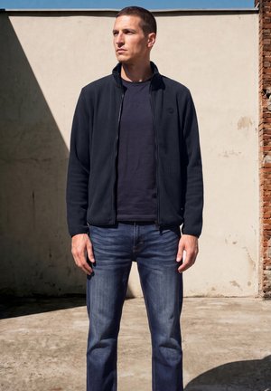 Homme portant une veste noire zippée, un t-shirt noir et un jean bleu, debout à l'extérieur contre un mur beige avec un bord en brique.