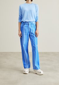 Maglione azzurro chiaro con maniche lunghe abbinato a pantaloni a gamba larga blu a righe. I pantaloni presentano una chiusura con bottone e tasche anteriori. Zoccoli bianchi.