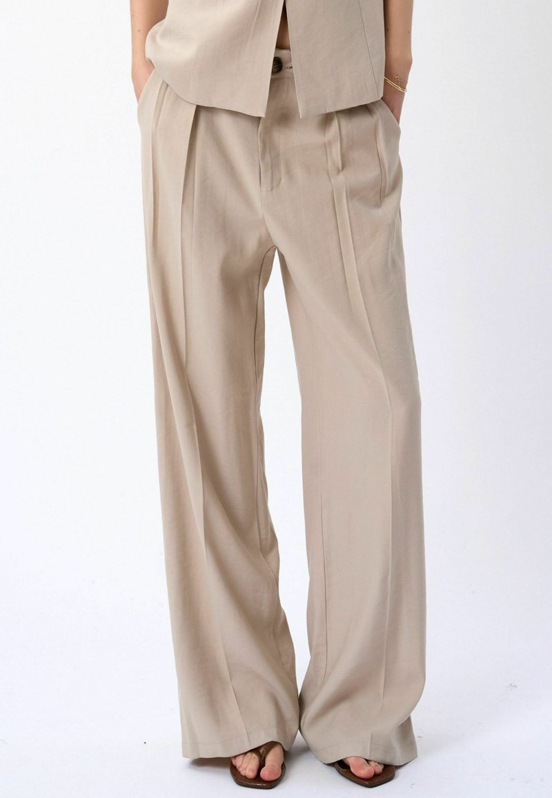 Pantalon beige à jambes larges avec plis, porté les mains dans les poches et sandales marron à bout ouvert sur fond blanc.