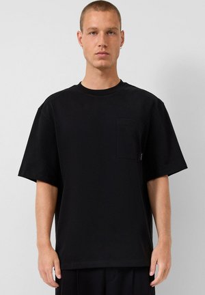 Młody mężczyzna o krótkich włosach, ubrany w prostą, czarną, oversize'ową koszulkę z kieszenią na piersi, stojący na tle jasnoszarego tła.