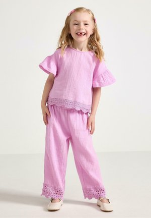 Usměvavá mladá dívka se světlými vlasy, oblečená v růžovém texturovaném topu a kalhotách s krajkovým lemováním a krémových botách, stojící na jednobarevném pozadí.