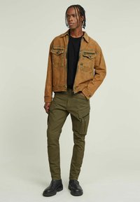 Giubbotto di jeans marrone con due tasche sul petto, t-shirt nera sotto, pantaloni cargo verde oliva con zip e stivali neri alla caviglia.