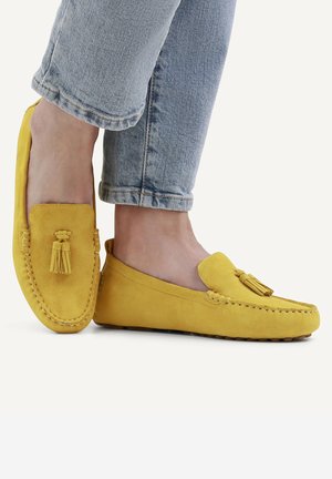 Füße in gelben Wildleder-Loafers mit Quasten, kombiniert mit hellblauen Denim-Jeans vor weißem Hintergrund.