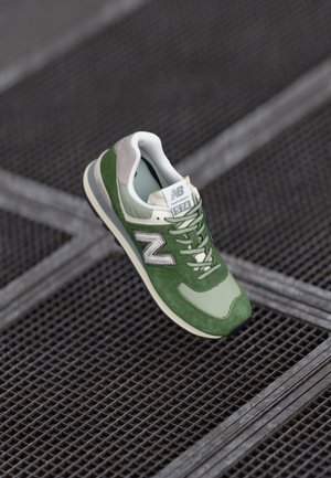 Grüner und beigefarbener New Balance 574 Sneaker, der über einem Metallgitterboden schwebt, mit sichtbaren detaillierten Schnürsenkeln und Logo.