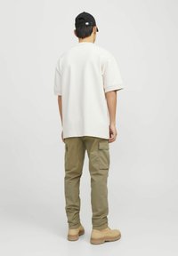 Übergro�ßes cremefarbenes T-Shirt, olivgrüne Cargo-Hosen und beige Stiefel, von hinten betrachtet. Eine schwarze Mütze rundet das Outfit ab. Schlichte Designelemente.