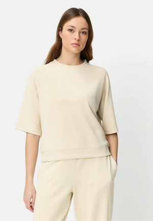 Femme aux longs cheveux bruns portant un sweat-shirt beige à manches courtes et un pantalon assorti, debout avec une main dans la poche.