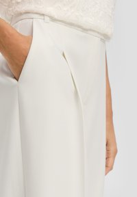 Białe dopasowane spodnie o gładkiej fakturze, z przeszyciami z przodu i bocznymi kieszeniami. Ręka umieszczona w lewej kieszeni.