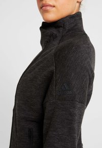 Černá zipová sportovní bunda z texturované tkaniny; má vysoký límec a tónové logo Adidas na levém rameni.