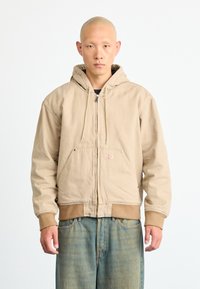 Beige hooded jacket with a zipper, two front pockets, and ribbed cuffs. Features a small logo on the chest and a relaxed fit design.