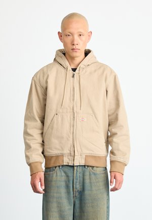 Beige hooded jacket with a zipper, two front pockets, and ribbed cuffs. Features a small logo on the chest and a relaxed fit design.