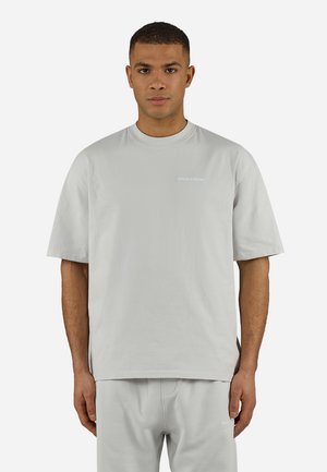 Ung mann iført en lys grå oversized T-skjorte med korte ermer og matchende bukser, stående foran en ensfarget hvit bakgrunn.