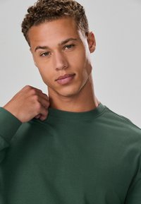 Green sweatshirt made of smooth fabric, featuring a crew neck and ribbed cuffs. Model has short curly hair and holds the sleeve with one hand.