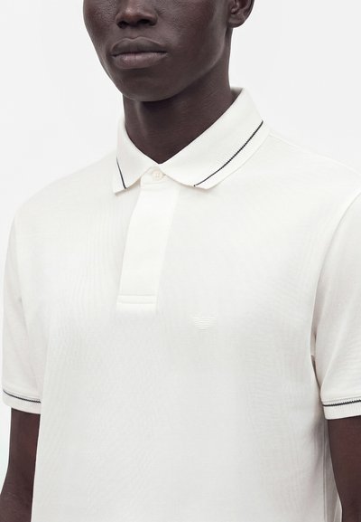 Homme portant un polo blanc avec des détails rayés noirs sur le col et les extrémités des manches, debout devant un fond uni.