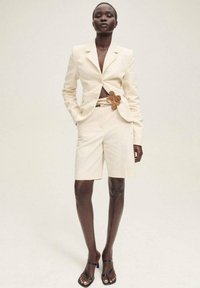 Blazer crème ajusté avec un design cintré, associé à un short assorti et une ceinture décorative à fleurs. Des sandales noires à lanières complètent le look.