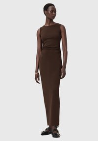 Brązowa, bezrękawnikowa sukienka maxi o dopasowanym kroju i gładkiej fakturze. Uzupełniona czarnymi butami z złotymi elementami ozdobnymi.