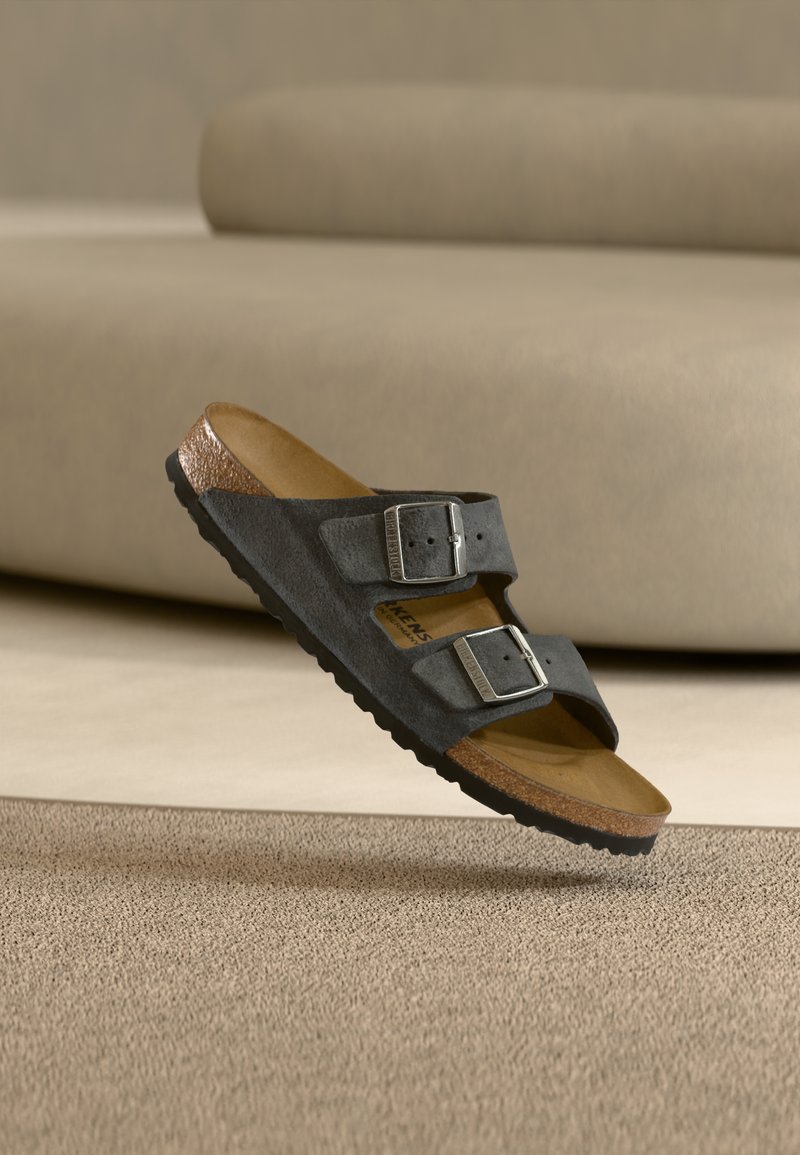 Σανδάλι από σκουρόχρωμο γκρίζο σουέτ με δύο ασημένιες αγκράφες και φελλό στη σόλα, εμφανιζόμενο αιωρούμενο πάνω από μπεζ υφασμάτινο χαλί και ουδέτερο φόντο.