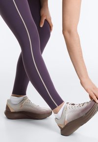 Personne portant des leggings violets et des chaussures de sport beige s'étire en touchant son pied avant, mettant l'accent sur les jambes et les mains.