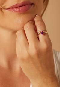 Drie ringen worden aan een hand gedragen: een gouden band met een paarse hartvormige edelsteen, een slanke gouden band en een band met kleine heldere stenen.