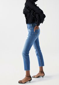 Jeans ajustados de color azul claro con detalles desgastados, combinados con una blusa negra con volantes y tacones negros con tiras, mostrando una vista lateral.