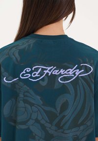 Teal T-shirt med et tonet grafisk design, der viser stiliseret skrifttype "Ed Hardy" i lavendel. Rund hals og korte ærmer.