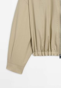 Beige jacket featuring a zip closure, elasticized hem, and wide sleeves. Made of lightweight fabric with a smooth texture.