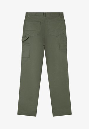 Mand stående iført hvid T-shirt med korte ærmer, olivengrønne bukser med knælapper, hvide sneakers og et perlearmbånd på højre håndled.