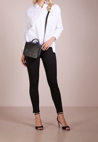 Schwarze Lederhandtasche mit geprägtem "HUGO"-Logo, kombiniert mit einem weißen Hemd, schwarzen Skinny-Jeans und schwarzen Riemchensandalen.