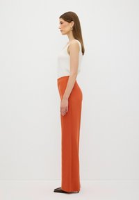 Witte geribde tanktop gecombineerd met hoogsluitende, strakke oranje broek. Model staat in profiel, met bruine puntige schoenen.