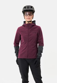 Burgunderfarbene Softshelljacke mit grauen Ärmeln, Frontreißverschluss und Kapuze; kombiniert mit schwarzen Hosen und Handschuhen. Getragen unter einem schwarzen Fahrradhelm und einer Brille.