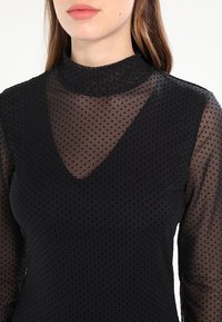 Schwarze Bluse aus transparentem gepunktetem Stoff, mit hohem Ausschnitt und V-förmiger Öffnung. Lange Ärmel und ein strukturierter Stoff sorgen für visuelles Interesse.