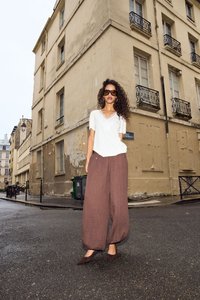 Femme aux cheveux bouclés portant des lunettes de soleil, un haut blanc et un pantalon marron ample, debout à un coin de rue mouillé près d’un vieux bâtiment beige.