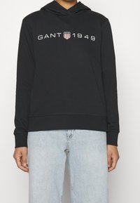 Černá mikina z bavlny, se předním logem "GANT 1949" a štítem. Dlouhé rukávy a volný střih. Nositá s světle modrými džíny.