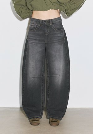 Personne portant un jean noir délavé taille haute à jambes larges et une veste courte verte olive avec des manches bouffantes, debout sur un sol uni.