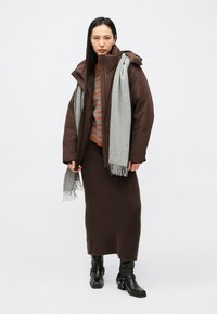 Brązowa puchowa kurtka, szary szalik, brązowo-pomarańczowy sweter w paski, brązowa spódnica, czarne botki. Gładkie tekstury, casualowy styl, warstwowy wygląd.