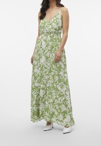 Maxi šaty v zelené látce s bílým květinovým vzorem, výstřih do V a seřiditelné tenké ramínka. Volný střih s definovaným pasem, doplněné bílými botami.