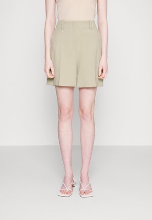 Vista de cintura a pies de una persona que lleva pantalones cortos plisados de tiro alto beige y sandalias blancas con tiras y tacón sobre un fondo liso.