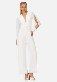 Tutina bianca con scollatura a V profonda, maniche lunghe in tessuto trasparente e vita arricciata. Design a gamba larga, presenta ritagli sulle spalle. Indossata con tacchi.
